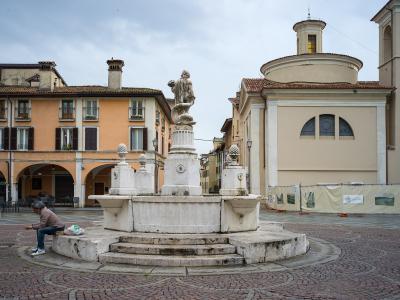 A stone fountain with a statue of a figure atop a column stands in a paved public square, flanked by an orange building with arched porticoes on the left and a beige church with a dome on the right; a person sits on the fountain’s edge.