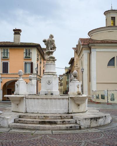A stone fountain with a statue of a figure atop a column stands in a paved public square, flanked by an orange building with arched porticoes on the left and a beige church with a dome on the right; a person sits on the fountain’s edge.