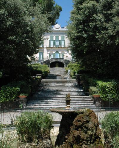 A view of a grand villa with green shutters and a long stone staircase leading up to it, framed by trees and featuring a fountain in the foreground.