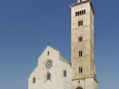 A low-angle photograph of a large, light-colored stone cathedral with a prominent square bell tower, a rose window, and an arched entrance bridge, set against a clear blue sky with the sea visible in the background.