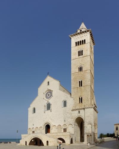 A low-angle photograph of a large, light-colored stone cathedral with a prominent square bell tower, a rose window, and an arched entrance bridge, set against a clear blue sky with the sea visible in the background.