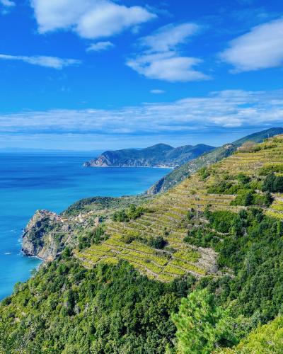 A scenic coastal landscape showing terraced vineyards on a steep hillside overlooking the blue sea, with a small village perched on a rocky promontory in the distance under a partly cloudy sky.