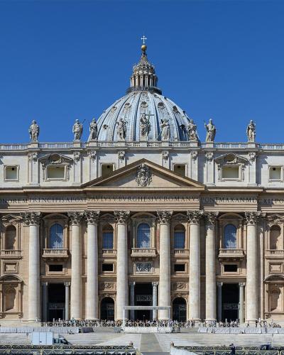 A frontal view of St. Peter's Basilica in Vatican City, showing its grand Baroque facade with Corinthian columns, statues along the roofline, the central dome by Michelangelo, and the Latin inscription "IN HONOREM PRINC APOST PAVLVS V BORGHESIVS R