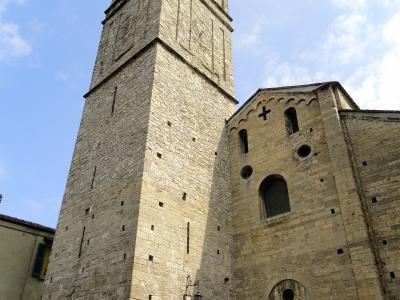 A low-angle photograph of a tall, square stone bell tower (campanile) attached to a Romanesque-style church facade with arched windows and a cross, under a blue sky with scattered clouds; a small plaque or sign is visible on the tower’s lower sect