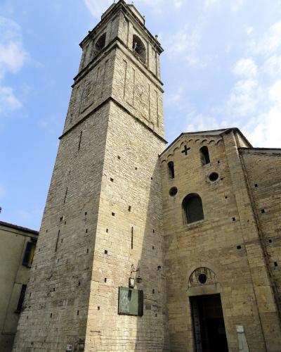 A low-angle photograph of a tall, square stone bell tower (campanile) attached to a Romanesque-style church facade with arched windows and a cross, under a blue sky with scattered clouds; a small plaque or sign is visible on the tower’s lower sect
