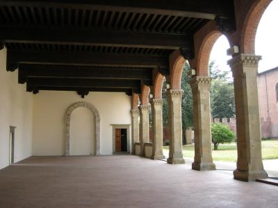 A covered loggia with brick arches, stone columns, and a dark wooden beam ceiling, featuring an arched doorway and a view of a garden and brick wall in the background.