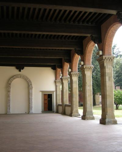 A covered loggia with brick arches, stone columns, and a dark wooden beam ceiling, featuring an arched doorway and a view of a garden and brick wall in the background.