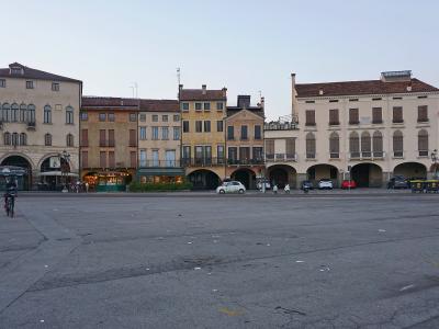 A wide, paved public square with a row of multi-story buildings featuring arched ground-floor arcades and shuttered windows, with a few cyclists and cars visible; no central island, canal, or ring of statues is present in the frame.