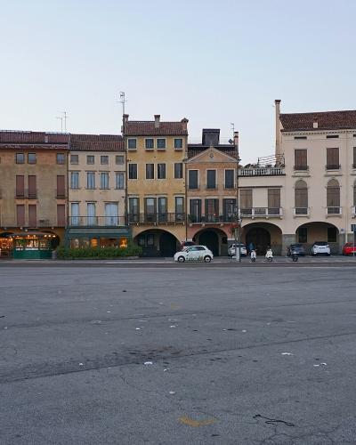 A wide, paved public square with a row of multi-story buildings featuring arched ground-floor arcades and shuttered windows, with a few cyclists and cars visible; no central island, canal, or ring of statues is present in the frame.