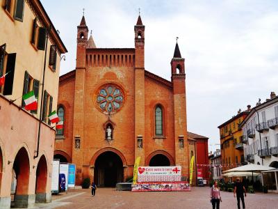 The image shows the red-brick facade of a large cathedral with a prominent rose window, two bell towers, and a central spire, situated in a paved town square with Italian flags and event banners.