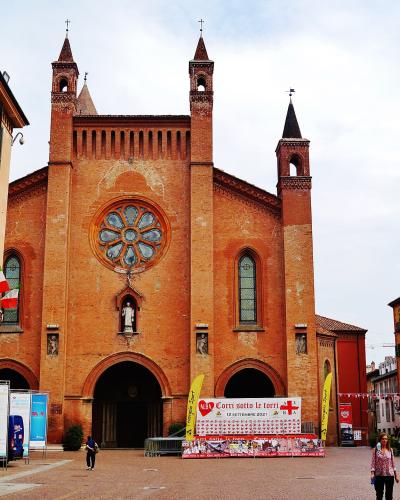 The image shows the red-brick facade of a large cathedral with a prominent rose window, two bell towers, and a central spire, situated in a paved town square with Italian flags and event banners.