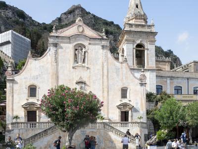 The image shows the facade of the Church of San Giuseppe in Taormina, Sicily, featuring its distinctive Baroque architecture, a bell tower, a statue in a niche, a flowering oleander tree in the foreground, and a checkered pavement plaza with touri