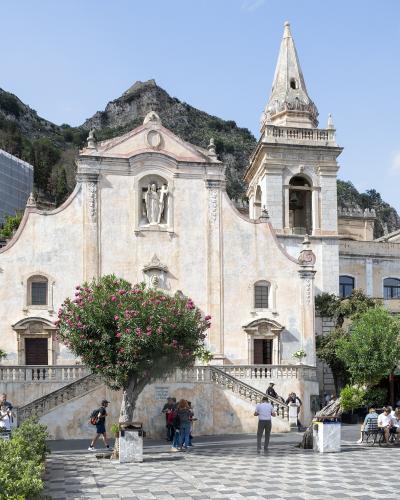 The image shows the facade of the Church of San Giuseppe in Taormina, Sicily, featuring its distinctive Baroque architecture, a bell tower, a statue in a niche, a flowering oleander tree in the foreground, and a checkered pavement plaza with touri