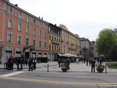 A wide-angle street-level view of a large, paved European town square lined with multi-story historic buildings in pastel colors (pink, yellow, beige), featuring outdoor café seating under umbrellas, pedestrians walking and gathering, planters, an