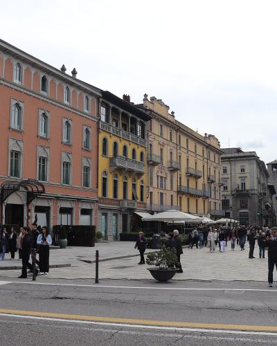 A wide-angle street-level view of a large, paved European town square lined with multi-story historic buildings in pastel colors (pink, yellow, beige), featuring outdoor café seating under umbrellas, pedestrians walking and gathering, planters, an