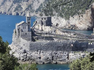 A stone fortress and church complex situated on a rocky promontory overlooking the sea, with a prominent bell tower and fortified walls, framed by foreground foliage.