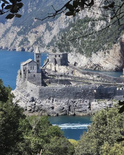A stone fortress and church complex situated on a rocky promontory overlooking the sea, with a prominent bell tower and fortified walls, framed by foreground foliage.