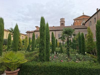 A formal garden with tall Italian cypress trees, rose bushes, and a potted palm in the foreground, set against a backdrop of historic brick buildings including a church with a bell tower and a cross.