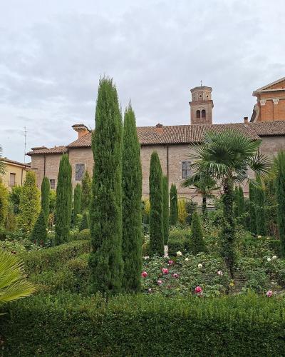 A formal garden with tall Italian cypress trees, rose bushes, and a potted palm in the foreground, set against a backdrop of historic brick buildings including a church with a bell tower and a cross.