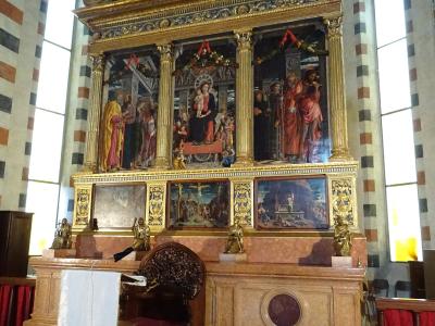 The image shows the ornate, gilded altarpiece and marble altar of a church interior, featuring religious paintings and sculptures, with distinctive striped stone columns and arched windows in the background.