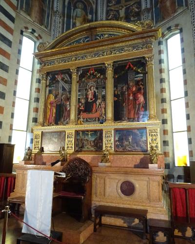 The image shows the ornate, gilded altarpiece and marble altar of a church interior, featuring religious paintings and sculptures, with distinctive striped stone columns and arched windows in the background.