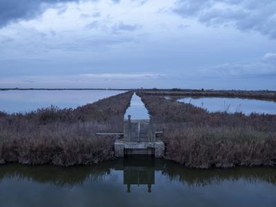 A long, straight canal or water channel flanked by grassy embankments and salt pans under a cloudy twilight sky, with a small concrete sluice gate structure in the foreground.