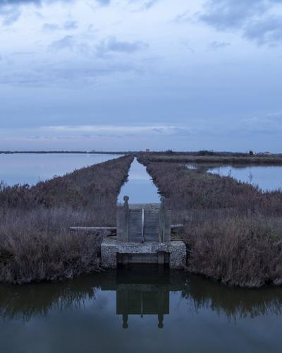 A long, straight canal or water channel flanked by grassy embankments and salt pans under a cloudy twilight sky, with a small concrete sluice gate structure in the foreground.