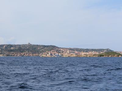 A coastal view of a town with terracotta-roofed buildings nestled along a rocky, hilly shoreline, viewed from the sea under a pale sky.