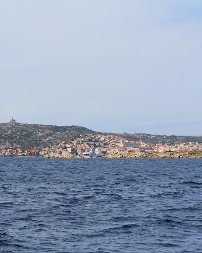A coastal view of a town with terracotta-roofed buildings nestled along a rocky, hilly shoreline, viewed from the sea under a pale sky.