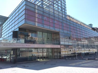 The image shows the modern glass-fronted exterior of a building with prominent signage reading "GALATA MUSEO DEL MARE" above the entrance and "GALATA" in large letters on the upper facade, situated in an open paved plaza under a clear sky.
