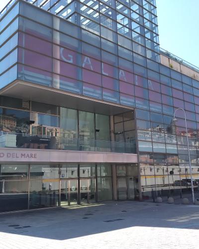 The image shows the modern glass-fronted exterior of a building with prominent signage reading "GALATA MUSEO DEL MARE" above the entrance and "GALATA" in large letters on the upper facade, situated in an open paved plaza under a clear sky.