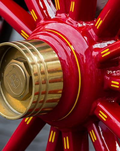 A close-up of a glossy red spoked wheel with yellow decorative stripes and a brass hubcap featuring a "B" logo, likely part of a carriage or coach.