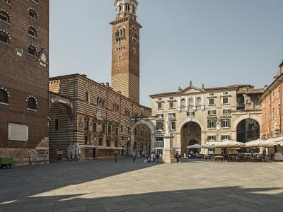 A wide-angle view of a large, paved public square in Verona, Italy, featuring the Palazzo della Ragione with its distinctive striped facade and the tall Torre dei Lamberti rising behind it, along with the Loggia del Consiglio and outdoor cafe seat