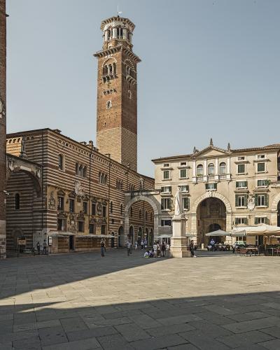 A wide-angle view of a large, paved public square in Verona, Italy, featuring the Palazzo della Ragione with its distinctive striped facade and the tall Torre dei Lamberti rising behind it, along with the Loggia del Consiglio and outdoor cafe seat