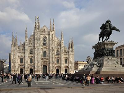 The image shows the facade of the Duomo di Milano cathedral in Milan, Italy, with the equestrian statue of Vittorio Emanuele II prominently in the foreground on the right, and crowds of tourists in the piazza.