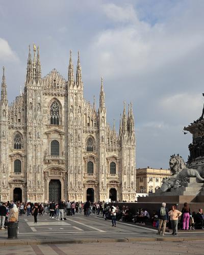 The image shows the facade of the Duomo di Milano cathedral in Milan, Italy, with the equestrian statue of Vittorio Emanuele II prominently in the foreground on the right, and crowds of tourists in the piazza.