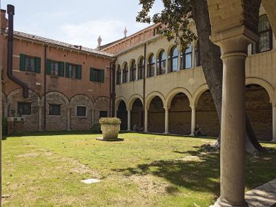 A sunlit courtyard with a grassy lawn, a central stone planter, and surrounding buildings featuring brick arches and a loggia with columns, identified by the filename as the cloister of the Church of Santa Maria della Visitazione (degli Artigianel