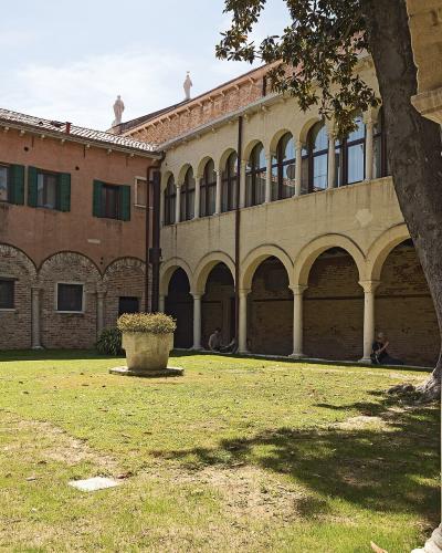 A sunlit courtyard with a grassy lawn, a central stone planter, and surrounding buildings featuring brick arches and a loggia with columns, identified by the filename as the cloister of the Church of Santa Maria della Visitazione (degli Artigianel
