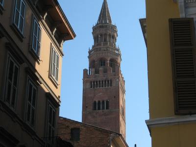 A tall, medieval brick bell tower with a pointed spire and golden orb finial, viewed from a narrow street flanked by buildings with shuttered windows under a clear blue sky.