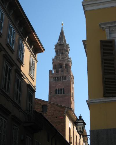 A tall, medieval brick bell tower with a pointed spire and golden orb finial, viewed from a narrow street flanked by buildings with shuttered windows under a clear blue sky.