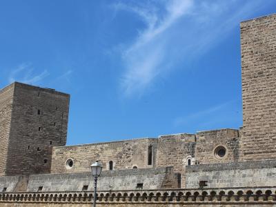 The image shows a large medieval stone fortress with two prominent square towers, crenellated walls, and circular windows, set against a blue sky with wispy clouds; a decorative balustrade and a lamppost are visible in the foreground.