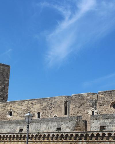 The image shows a large medieval stone fortress with two prominent square towers, crenellated walls, and circular windows, set against a blue sky with wispy clouds; a decorative balustrade and a lamppost are visible in the foreground.