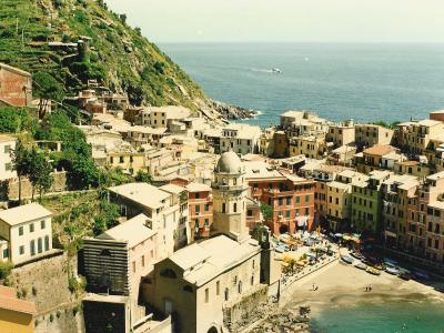 A high-angle view of the coastal village of Vernazza in Italy, showing the Church of Santa Margherita d'Antiochia, the small harbor with boats, colorful buildings cascading down the hillside, and the Ligurian Sea.