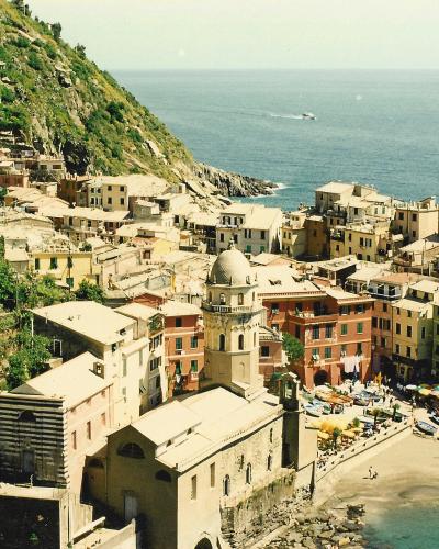 A high-angle view of the coastal village of Vernazza in Italy, showing the Church of Santa Margherita d'Antiochia, the small harbor with boats, colorful buildings cascading down the hillside, and the Ligurian Sea.