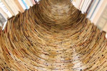 A close-up, low-angle photograph looking up into a spiral tunnel or tower constructed from the pages and spines of numerous books, with visible text on some pages and book edges.