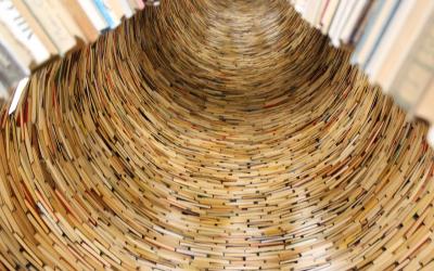 A close-up, low-angle photograph looking up into a spiral tunnel or tower constructed from the pages and spines of numerous books, with visible text on some pages and book edges.