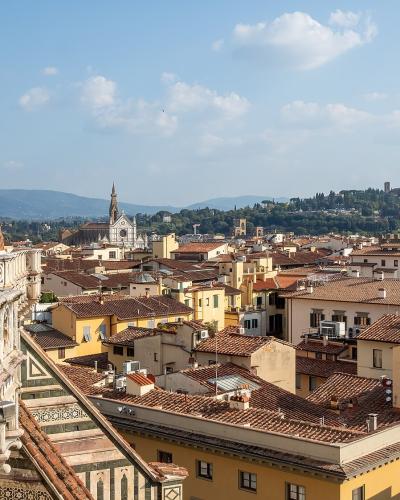 A high-angle view from the Duomo terrace showing the terracotta dome of the Florence Cathedral on the left, overlooking the city's rooftops with the Palazzo Vecchio tower and Giotto's Campanile visible in the distance.