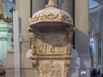 An ornate, multi-tiered marble holy water font (acquasantiera) topped with a statue of the Virgin Mary, situated indoors between large stone columns within a cathedral.