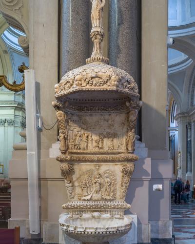 An ornate, multi-tiered marble holy water font (acquasantiera) topped with a statue of the Virgin Mary, situated indoors between large stone columns within a cathedral.
