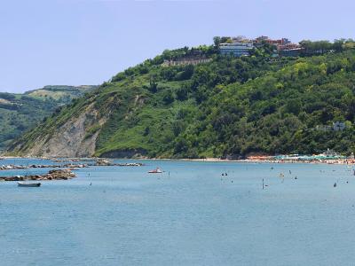 A coastal scene showing a sandy beach crowded with people, turquoise water, and a steep, green hillside with buildings, including a prominent white hotel, rising above the shore.